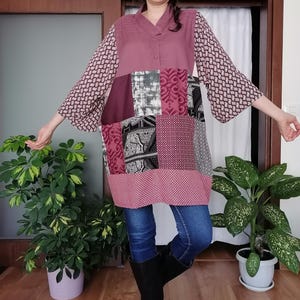 May include: A patchwork tunic in shades of mauve, burgundy, grey, and white. The tunic features a V-neck, long sleeves with a geometric pattern, and a mix of patterned and solid fabric panels. The person is wearing blue jeans and black boots.