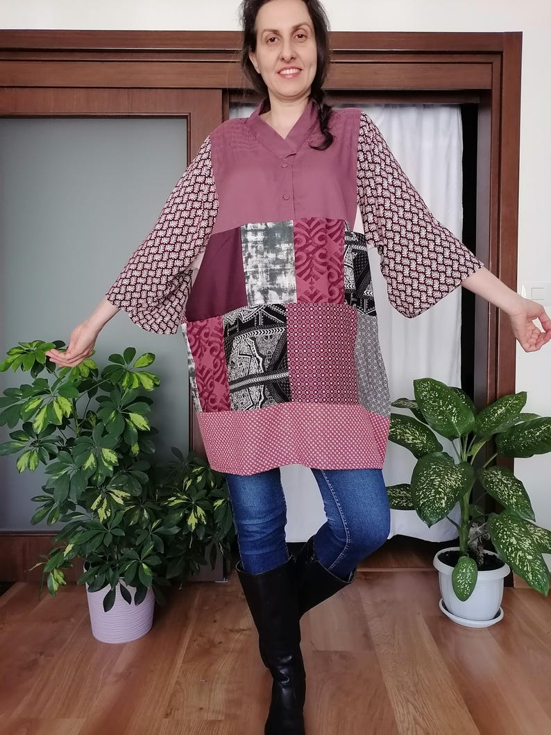 May include: A patchwork tunic in shades of mauve, burgundy, grey, and white. The tunic features a V-neck, long sleeves with a geometric pattern, and a mix of patterned and solid fabric panels. The person is wearing blue jeans and black boots.