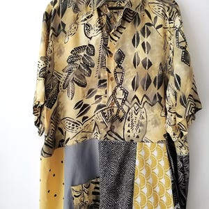 May include: A short-sleeved button-down shirt with a patchwork design. The top portion features a beige and black leaf pattern. The bottom is a patchwork of yellow, grey, and black fabrics with various patterns. The shirt is hanging on a wooden hanger.