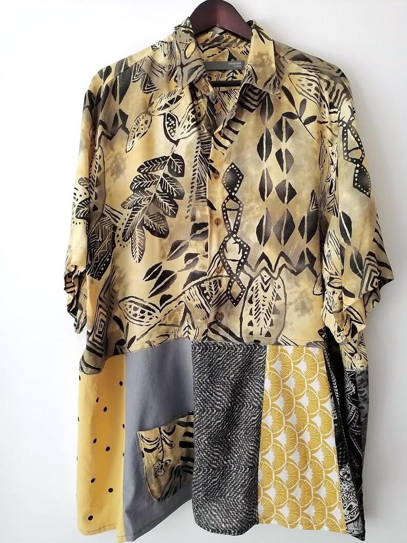 May include: A short-sleeved button-down shirt with a patchwork design. The top portion features a beige and black leaf pattern. The bottom is a patchwork of yellow, grey, and black fabrics with various patterns. The shirt is hanging on a wooden hanger.