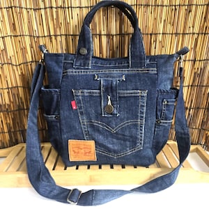 May include: A blue denim tote bag with a shoulder strap. The bag has multiple pockets, a button closure, and a brown leather patch with red text. The bag is made from denim fabric and has a classic design.