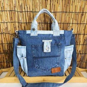 Może przedstawiać: Dżinsowa torba typu tote z jasnoniebieskim dżinsowym uchwytem i paskiem na ramię. Torba posiada dużą przednią kieszeń i dwie boczne kieszenie, wszystkie wykonane z przetworzonych dżinsów. Mała brązowa skórzana metka jest przyszyta z przodu.