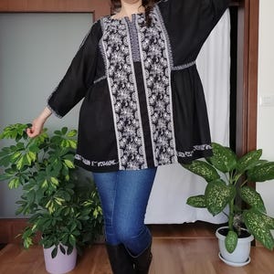 May include: Black tunic top with white floral embroidery. The top has long sleeves and a round neckline. The embroidery is detailed and covers the front and sleeves. The tunic is paired with blue jeans and black boots.