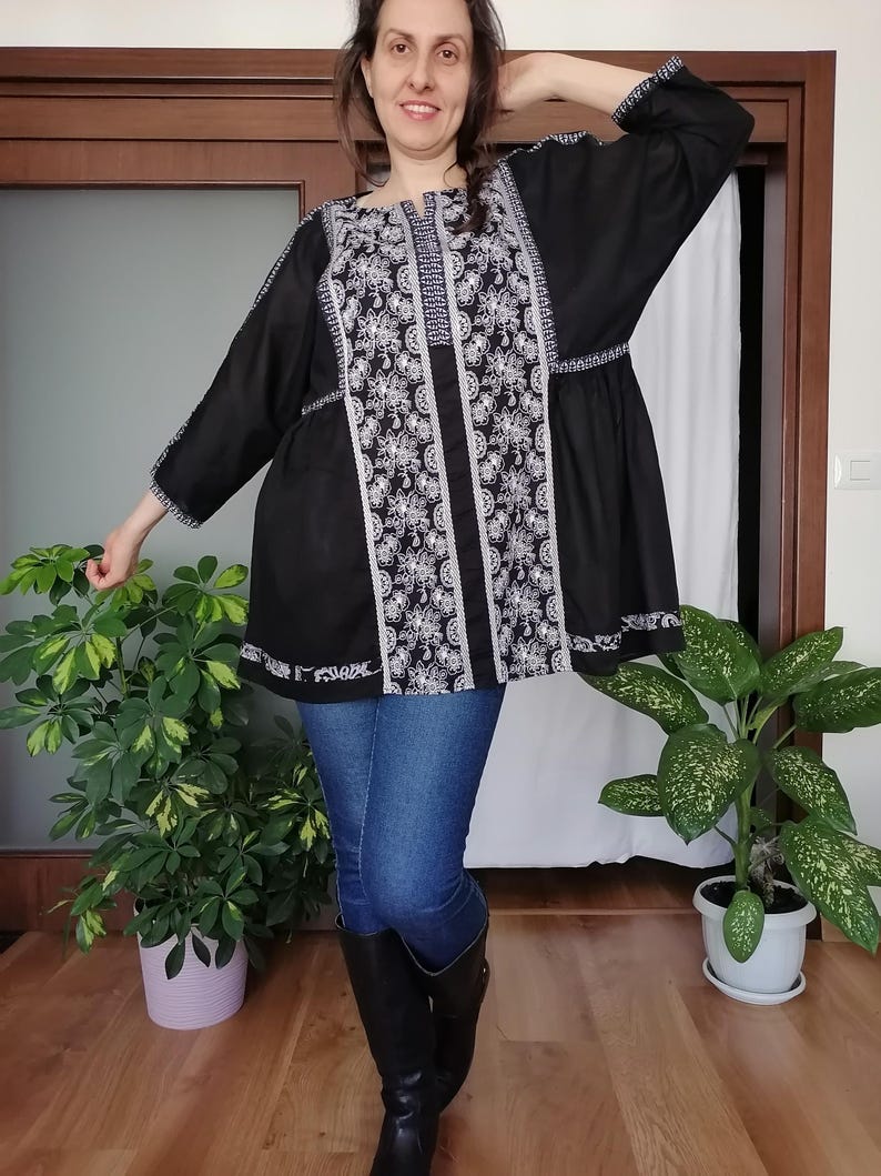May include: Black tunic top with white floral embroidery. The top has long sleeves and a round neckline. The embroidery is detailed and covers the front and sleeves. The tunic is paired with blue jeans and black boots.