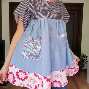 May include: A patchwork tunic dress with a striped top, light blue denim-style fabric, and floral details. The dress has a pink floral hem and pocket details. It features short sleeves and a button-down front.