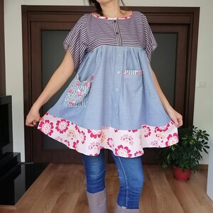 May include: A handmade tunic dress with a striped top, light blue body, and floral trim. The dress features a button-down front, two pockets, and a pink floral pattern on the hem. The sleeves are short and the dress is knee-length.