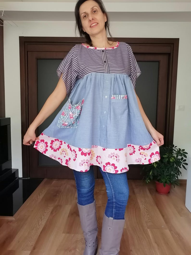 May include: A handmade tunic dress with a striped top, light blue body, and floral trim. The dress features a button-down front, two pockets, and a pink floral pattern on the hem. The sleeves are short and the dress is knee-length.