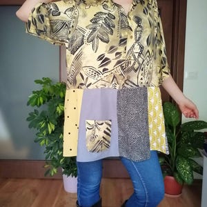 May include: A patchwork tunic shirt with a yellow and black patterned top, a light purple pocket, and various fabric panels in yellow, grey, and black. The shirt is worn with blue jeans and black boots.