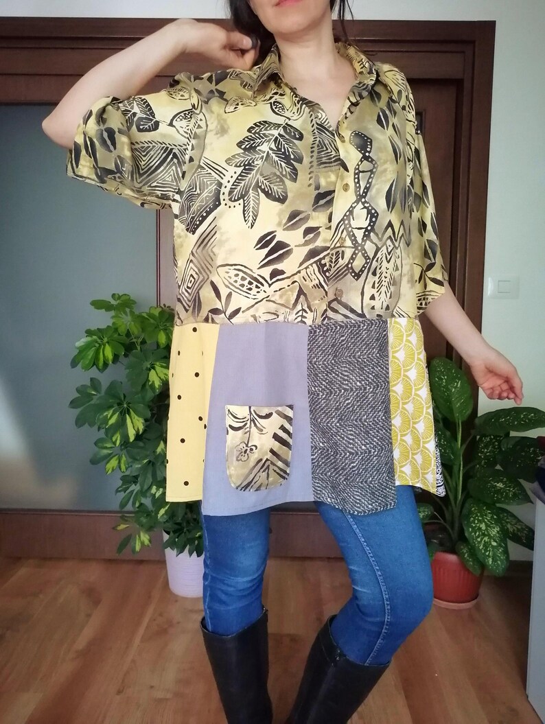 May include: A patchwork tunic shirt with a yellow and black patterned top, a light purple pocket, and various fabric panels in yellow, grey, and black. The shirt is worn with blue jeans and black boots.