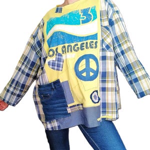 Op de afbeelding: Een geel overhemd met lange mouwen met een blauw en wit vredesteken, een blauw hart en de tekst "Los Angeles" gedrukt op de voorkant. Het overhemd is gelaagd over een blauw en geel geruit overhemd.