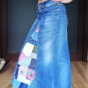 Op de afbeelding: Een lange blauwe spijkerrok met een voorpaneel opgebouwd uit een patchwork van diverse stoffen, waaronder bloemenprints, effen kleuren en denimresten. De rok heeft een klassiek ontwerp met vijf zakken en een knoop- en ritssluiting.