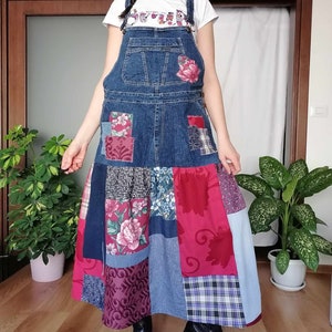 Peut inclure: Une robe salopette en jean avec des détails patchwork. La robe présente un haut à bavette, des poches et une longue jupe avec divers motifs de tissu, notamment des motifs floraux et à carreaux. La robe est portée avec un t-shirt blanc et des bottes noires.