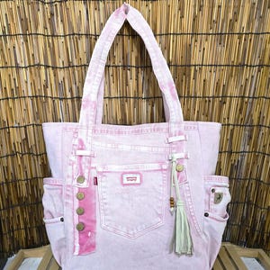 Puede incluir: Un bolso tote de mezclilla rosa claro con un bolsillo central con etiqueta de la marca Levi's, dos correas para el hombro y bolsillos laterales. El bolso está decorado con una fila vertical de cuatro botones metálicos, una etiqueta roja y una borla.