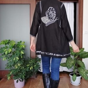 May include: Black tunic featuring a white embroidered snail design on the back. The tunic has three-quarter sleeves and a decorative white trim. The person is wearing blue jeans and black boots.