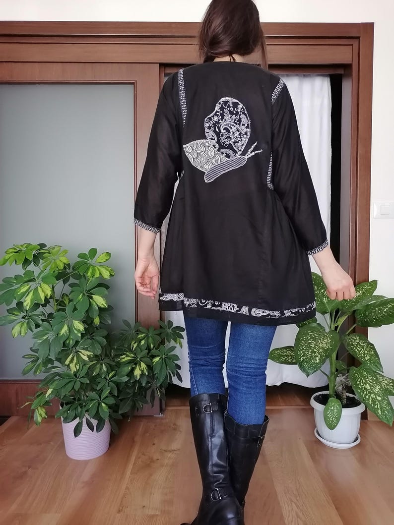 May include: Black tunic featuring a white embroidered snail design on the back. The tunic has three-quarter sleeves and a decorative white trim. The person is wearing blue jeans and black boots.