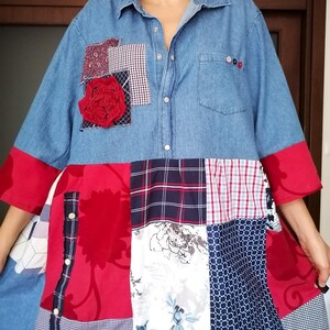 Può includere: Una camicia di jeans blu con un design a patchwork. La camicia ha un'applicazione di un fiore rosso, una tasca con bottoni e un motivo a quadri sul fondo.