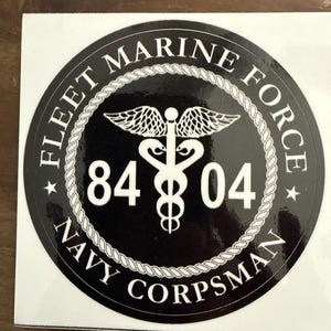Op de afbeelding: Een zwart-witte sticker met de tekst "FLEET MARINE FORCE NAVY CORPSMAN" rond een centraal embleem. Het embleem bevat een caduceussymbool met vleugels en de cijfers "84 04". Het ontwerp is omlijst door een touwachtig patroon.