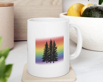 May include: White ceramic mug featuring a silhouette of three pine trees against a rainbow gradient background. The mug has a curved handle and sits on a beige coaster. The design is simple and graphic, suitable for hot beverages.