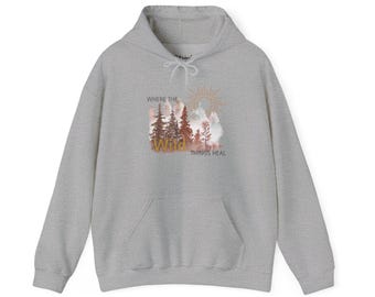 May include: A light gray hoodie with a front pocket and a drawstring hood. The front of the hoodie features a graphic of a forest scene with the text "WHERE THE WILD THINGS HEAL" in a stylized font. The graphic includes shades of brown, orange, and white.