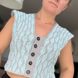 Puede incluir: Un chaleco de ganchillo sin mangas con un patrón de hojas de color azul claro y ribetes grises. La prenda tiene un escote en V y una tapeta vertical central abrochada con cinco botones negros redondos con un patrón de punto texturizado.