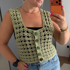 May include: A sage green sleeveless crochet vest featuring an openwork shell stitch pattern, a square neckline, and a front button placket with five matching round green buttons. The garment is cropped in length and has a textured, lightweight construction.