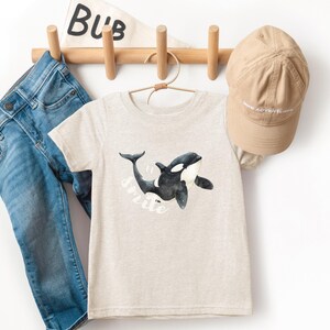 Könnte beinhalten: Ein hellbeige T-Shirt mit einer Aquarellillustration eines schwarz-weißen Orca-Wals und dem Wort "smile". Das Shirt hängt mit einer blauen Jeans und einer beigefarbenen Baseballkappe.