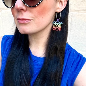May include: A pair of hoop earrings featuring a rainbow-colored sea turtle design. The earrings are suspended from thin, silver-colored hoops. The turtle design has a gradient of colors, including pink, orange, yellow, green, and blue.
