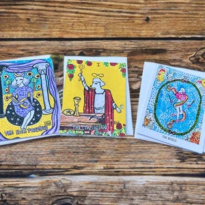 May include: Three tarot cards with colorful illustrations and text. The cards are "The High Priestess", "The Magician", and "The World". The cards are on a wooden surface. The illustrations feature figures, symbols, and vibrant colors.