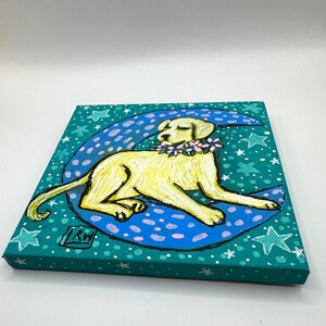 May include: A yellow Labrador Retriever dog with a pink flower garland around its neck is lying on a blue crescent moon with white and pink polka dots. The background is a green canvas with white stars.