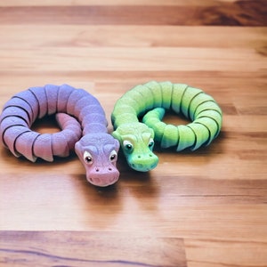 May include: Two colorful toy snakes, one purple and one green, are curled up on a wooden surface. The snakes have realistic features, including eyes and scales.