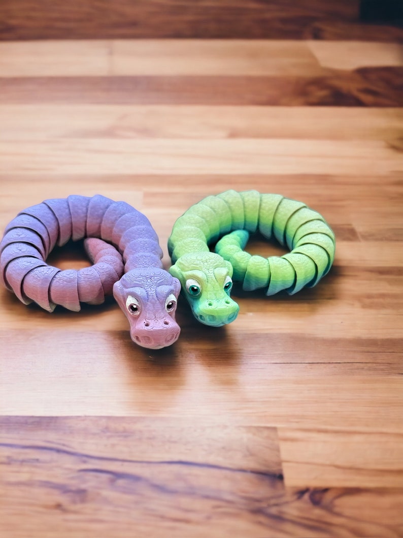May include: Two colorful toy snakes, one purple and one green, are curled up on a wooden surface. The snakes have realistic features, including eyes and scales.