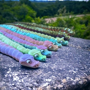 May include: A row of colorful, segmented, toy snakes with googly eyes. The snakes are arranged in a line on a gray, textured surface. The snakes are made of a soft, pliable material and are perfect for imaginative play.
