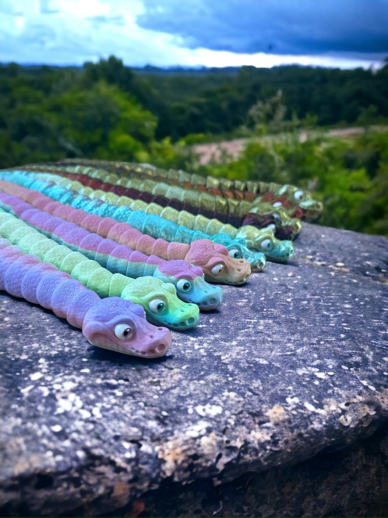 May include: A row of colorful, segmented, toy snakes with googly eyes. The snakes are arranged in a line on a gray, textured surface. The snakes are made of a soft, pliable material and are perfect for imaginative play.