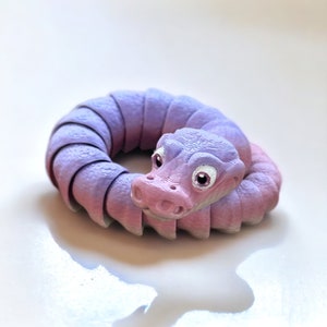 May include: A purple and pink segmented snake toy with a cute face and large white eyes. The snake is curled into a circle.