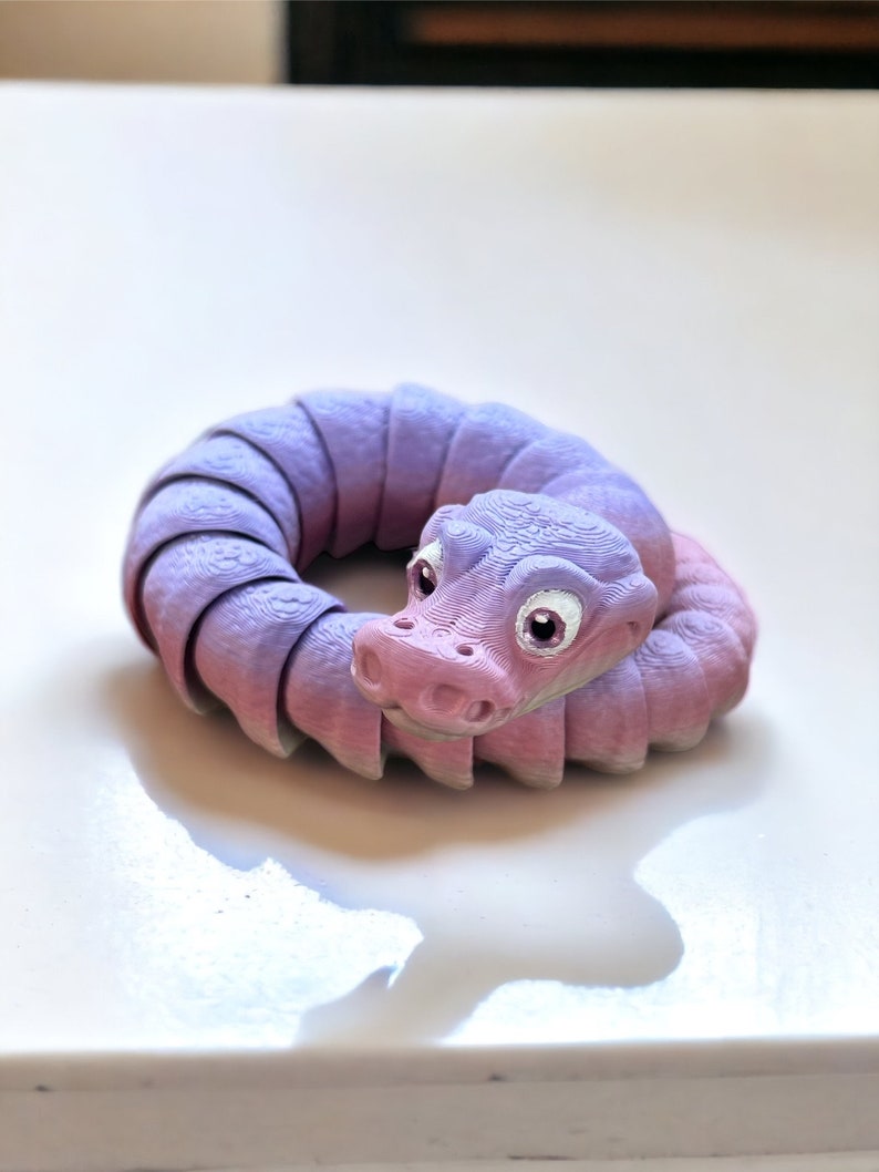 May include: A purple and pink segmented snake toy with a cute face and large white eyes. The snake is curled into a circle.