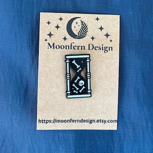 May include: Black and white hourglass pin with a skull and crossbones design. The pin is on a brown card with the text "Moonfern Design" and a website address. The background is a blue fabric.