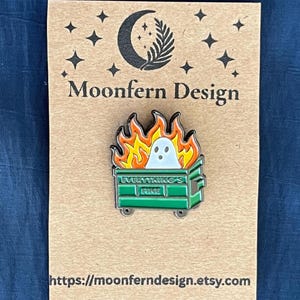 May include: Enamel pin featuring a ghost in a dumpster engulfed in flames. The green dumpster has the words "Everything's Fine" on it. The pin is on a brown card with the text "Moonfern Design" and a website address.