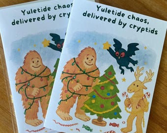 May include: Two Christmas cards with illustrations of a Bigfoot-like creature wrapped in Christmas lights, a Christmas tree, and a reindeer holding candy canes. The cards have the text "Yuletide chaos delivered by cry" and "Yuletide chaos, delivered by cryptids".