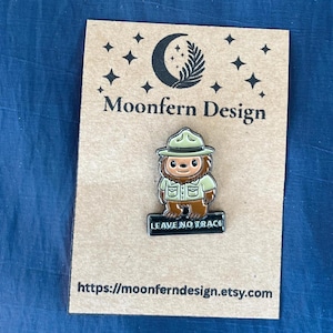 May include: Enamel pin featuring a cartoon Bigfoot character in a park ranger uniform, with the text "LEAVE NO TRACE". The pin is on a brown card with the text "Moonfern Design" and a website address.