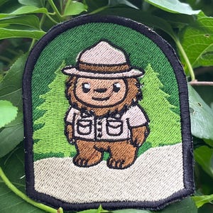 May include: An embroidered patch featuring a brown, furry creature wearing a tan park ranger hat and a light-colored button-down shirt with pockets. The character stands on a light-colored path against a green background with pine tree silhouettes.