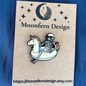 May include: A pin featuring a skeleton wearing sunglasses, riding a unicorn pool float with a rainbow mane and tail. The pin is on a brown card with the text "Moonfern Design" and a website address.