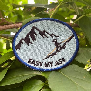 May include: A circular embroidered patch with a light blue background and a dark blue border. It features a black mountain silhouette and a black figure skiing down a slope. The text "EASY MY ASS" is embroidered in black at the bottom.