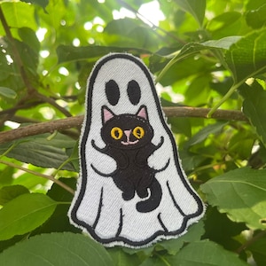 May include: An embroidered patch featuring a white ghost with black outlines and two oval eyes. The ghost is holding a small black cat with large yellow eyes, pink inner ears, and a small pink nose. The design is rendered in thread on a white fabric base.