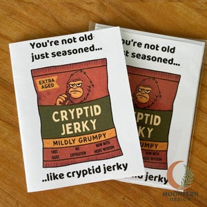 May include: Two greeting cards with the text "You're not old just seasoned..." and a graphic of a Cryptid Jerky bag. The bag is brown and green with a grumpy ape-like creature. The cards are on a wooden surface.