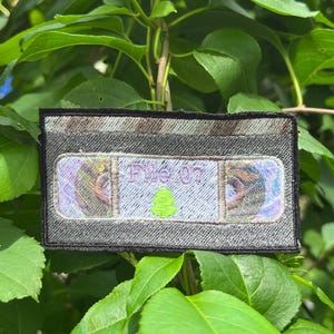 May include: A rectangular embroidered patch shaped like a VHS cassette tape. The patch features a dark gray border, a silver top section, and a central label area with iridescent accents. The text "File 07" is stitched in purple above a small green icon.