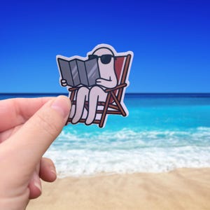 Może przedstawiać: Naklejka przedstawiająca postać z kreskówki w okularach przeciwsłonecznych czytającą mapę, siedzącą na krześle plażowym. Postać jest biała z czarnymi okularami przeciwsłonecznymi. Krzesło plażowe jest brązowe z czerwonym oparciem.