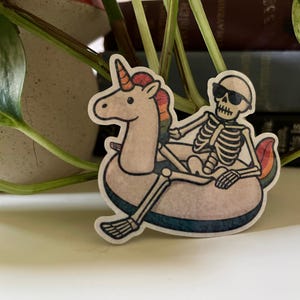Può includere: Un adesivo fustellato raffigurante uno scheletro beige con occhiali da sole neri, seduto all'interno di un salvagente a forma di unicorno beige. L'unicorno ha criniera e coda arcobaleno, e il salvagente ha un bordo inferiore blu scuro.
