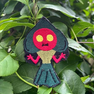 May include: An embroidered patch featuring a stylized character with a spade-shaped head, a bright red face, and two yellow circular eyes. The character has a tan torso, pink arms with three-fingered hands, and a dark green skirt-like lower body.