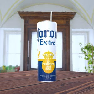 Puede incluir: Un vaso de acero inoxidable blanco y azul con un diseño de lata de cerveza Corona Extra. El vaso tiene una pajita y dice "La Cerveza Mas Fina" y "Imported Beer From Mexico 20 fl. oz."