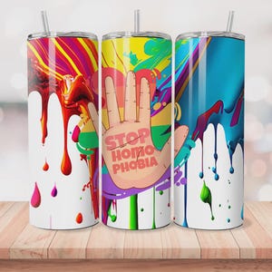 Puede incluir: Tres vasos de acero inoxidable con diseños abstractos coloridos y pajitas. Un vaso presenta una mano con el texto "STOP HOMOPHOBIA" en un esquema de color arcoíris. Los otros dos vasos tienen diseños de goteo de pintura.